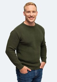 Dunkelgrüner, gerippter Strickpullover mit rundem Ausschnitt und langen Ärmeln, kombiniert mit blauen Denim-Jeans. Das Model posiert mit Händen in den Taschen.
