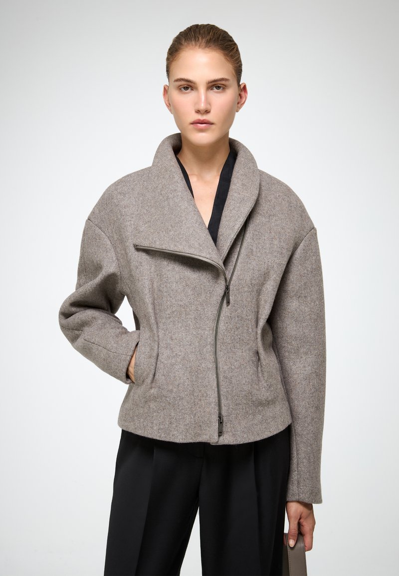 Sivi vuna jakna s visokim ovratnikom, asimetričnim zatvaračem i blago prevelikim krojem. Glatka tekstura, minimalistički dizajn.