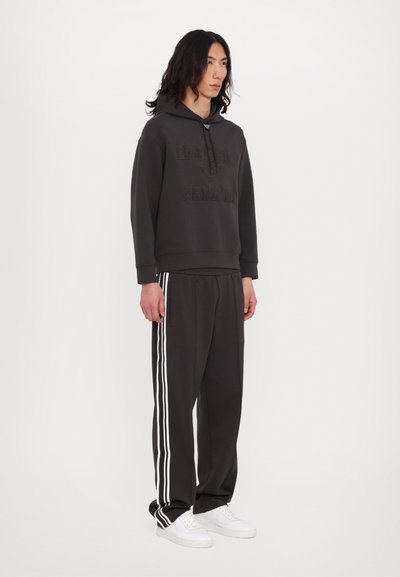 Corta escura com capuz e logótipo em relevo "Emporio Armani", combinada com calças de fato correspondentes com três riscas brancas nas laterais. Ténis brancos.