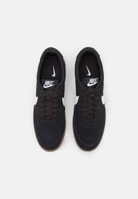 Fekete velúr sportcipők, textúrált felülettel, fehér Nike logóval, fűzős kialakítással és goma gumi talppal.