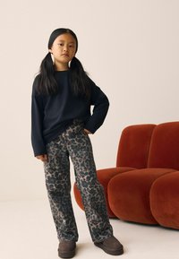 Sort langærmet skjorte, dyreprint brede bukser i grå og brune nuancer, brune slip-on sko. Baggrunden har en orange sofa.