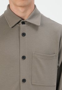 Tan button-up shirt with a textured fabric, featuring a collar, three black buttons, and a pocket on the left chest.