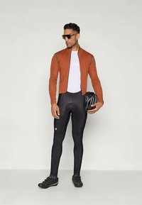 Velosipēdu apģērbs ar rūsas krāsas, ar rāvējslēdzēju aizveramu jaku un pielāgotu baltu pamatuslāni; melnām, elastīgām legingām; brillēm; un velosipēda ķiveri.