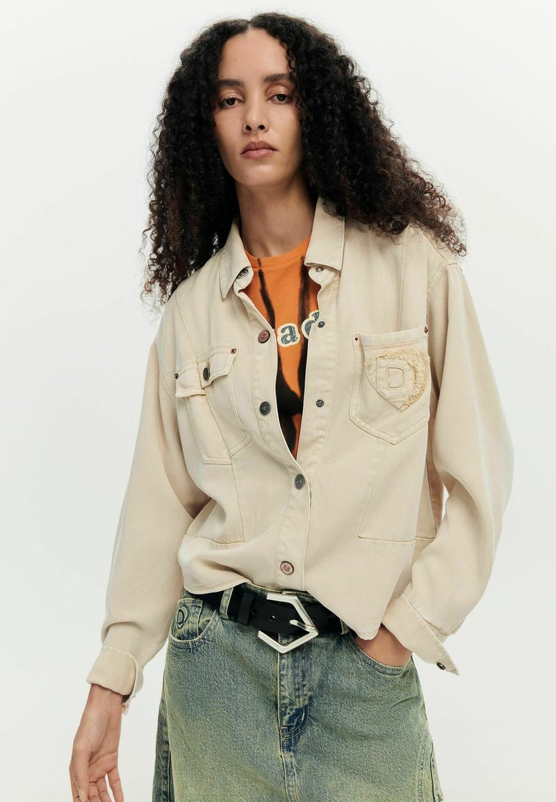 Femme aux cheveux bouclés portant une chemise beige boutonnée par-dessus un t-shirt graphique orange, rentrée dans un jean bleu délavé avec une ceinture noire.
