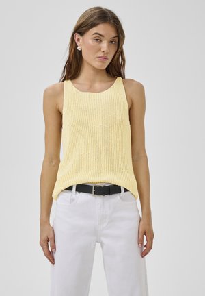 Mladá žena v žltom pletenom topu bez rukávov, biele nohavice s čiernym opaskom a perlovými náušnicami, stojaca pred jednoduchým pozadím.