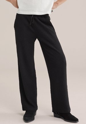 Person trägt schwarze, strukturierte Hosen mit weitem Bein und Tunnelzug an der Taille, kombiniert mit schwarzen Stiefeletten und einem weißen Oberteil.