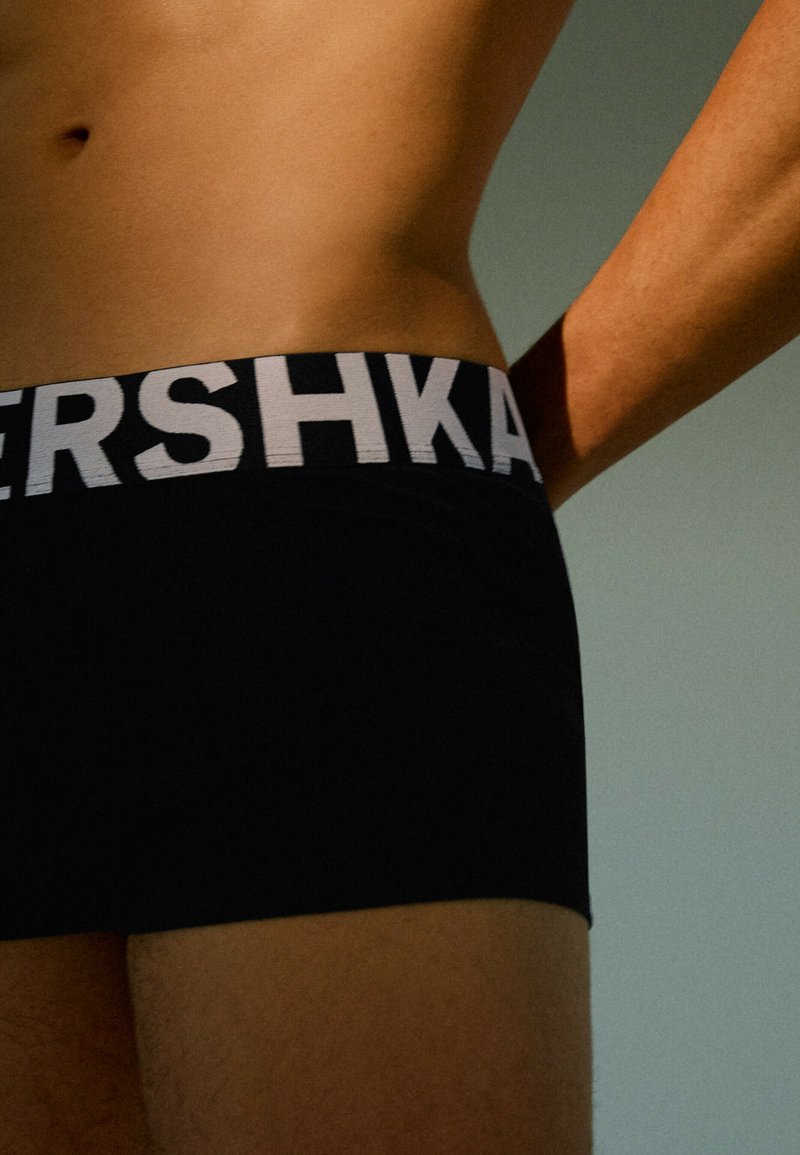 Torse masculin portant un boxer noir avec une ceinture élastique blanche présentant un texte partiellement en gras sur un fond uni atténué.