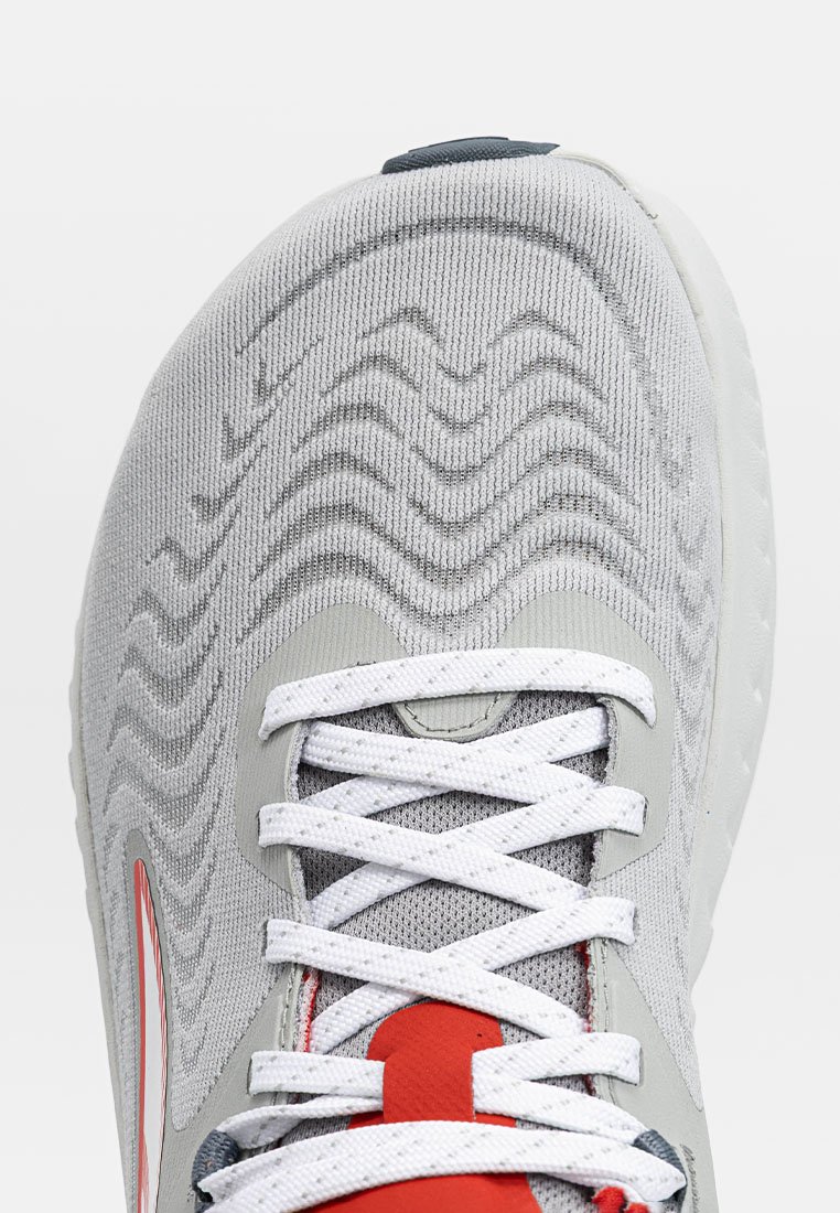 Zapatilla deportiva de color gris claro con parte superior de tela texturizada, cordones blancos y detalles rojos. Presenta una forma curva y un collar acolchado para mayor comodidad.