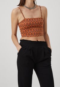 Haut court orange avec un motif ondulé marron ; présente des bretelles fines et un décolleté carré. Associé à un pantalon taille haute noir.