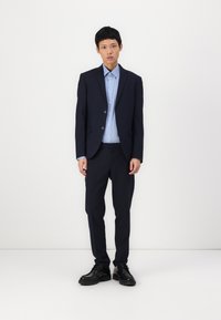 Costum cu dungi navy, cu o jachetă confecționată pe măsură și pantaloni asortați, purtat cu o cămașă ușor albastră cu nasturi și pantofi negri.