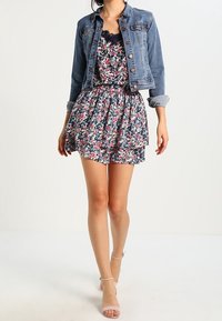 Dżinsowa kurtka nałożona na sukienkę w kwiatowym wzorze z falbanką. Sukienka ma odcienie granatu, różu i bieli, zestawiona z przezroczystymi obcasami.