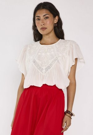 Femme aux longs cheveux foncés porte un chemisier blanc brodé à manches courtes et une jupe rouge taille haute, debout devant un fond uni.