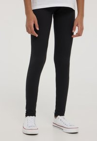 Czarne legginsy wykonane z elastycznego materiału, dopasowany krój, pełna długość do kostek, w połączeniu z białymi sneakersami z czerwonym akcentem.