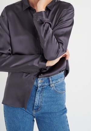 Persona con una camisa satinada de manga larga oscura abotonada, metida dentro de unos vaqueros azules de talle alto, con los brazos cruzados y una mano tocando la barbilla.