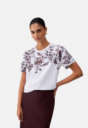 Donna con capelli scuri, indossa una camicia bianca a maniche corte con stampa floreale e una gonna scura, in piedi con una mano sul fianco davanti a uno sfondo semplice.