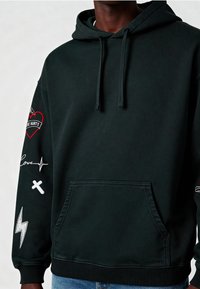 Felpa nera con tasca frontale, cordini e dettagli ricamati: un cuore, una linea del battito cardiaco, una croce e un fulmine sulle maniche.