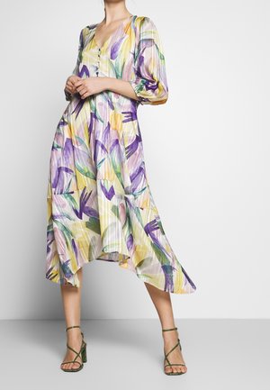 Femme portant une robe midi fluide à fleurs avec manches bouffantes et boutons sur le devant, associée à des talons verts à brides.