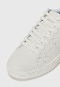 Sneaker en cuir blanc avec un embout rond, surface texturée, lacets blancs plats et détails de couture subtils. Design et couleur minimalistes.