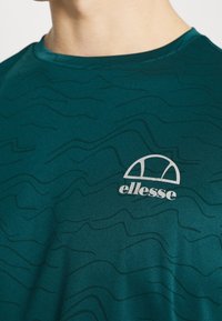 Turkusowa sportowa koszulka z wzorem fal, wykonana z gładkiej tkaniny. Małe białe logo po lewej stronie klatki piersiowej. Okrągły dekolt.