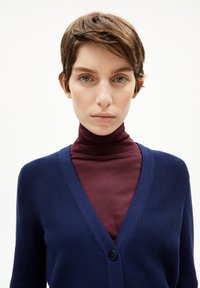 Dunkelblauer Cardigan mit strukturiertem Strick, ausgestattet mit einem V-Ausschnitt und einer Knopfverschluss, über einem enganliegenden burgunderfarbenen Rollkragenpullover getragen.
