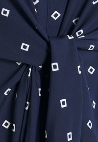 Navy sininen kangas, jossa on valkoisia neliökuvioita, sidottuna suureen löysään solmuun, näyttäen pehmeitä taitoksia ja rakennetta.