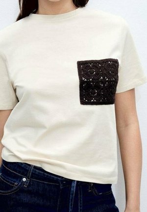 Kremowy t-shirt z krótkim rękawem z fakturowaną czarną kieszenią z szydełka. Prosty okrągły dekolt i luźny krój; na wysokości talii widoczne dżinsy.
