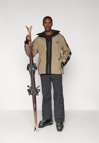 Χειμερινή στολή σκι με ταμπά μπουφάν με μαύρες λεπτομέρειες, μαύρο παντελόνι και μπότες σκι, κρατώντας πέδιλα σκι με δεσίματα. Απλό φόντο.