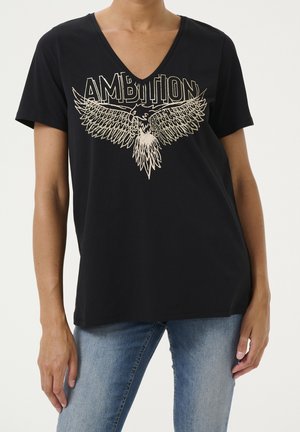 Kvinde iført sort V-hals T-shirt med guld ørne grafik og ordet "AMBITION," kombineret med blå jeans.