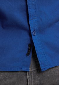 Modrá košile na knoflíky vyrobená z texturované bavlny, s postranní etiketou a rovnou spodní částí. Nositá přes šedé džíny.