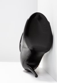 Chaussure à talon noir montrant une semelle texturée avec le logo "GUESS", positionnée à la verticale contre un fond d'angle blanc.