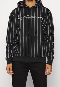 Mustapaita, jossa on valkoiset pystysuorat raitakuviot ja valkoinen käsinkirjoitettu logo rinnan kohdalla, henkilö, jolla kädet rentoina sivuilla.