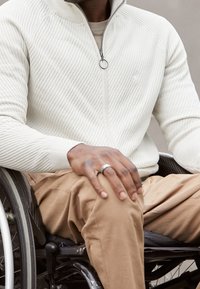 Bílý žebrovaný svetr na zip s vysokým límcem, šedé tričko, a béžové kalhoty, oblečené osobou sedící v invalidním vozíku. Stříbrný prsten na ruce.