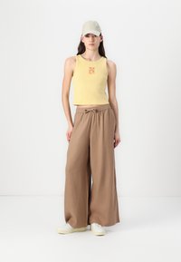 Yellow ribbed crop top with orange graphic, brown wide-leg pants, and beige cap; model wears white sneakers with a textured sole.