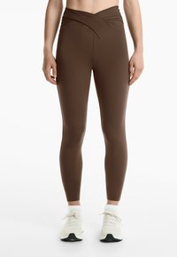 Bruna högmidjade leggings med en vriden design vid midjan. Tillverkade av en elastisk tygkvalitet, de passar nära kroppen.