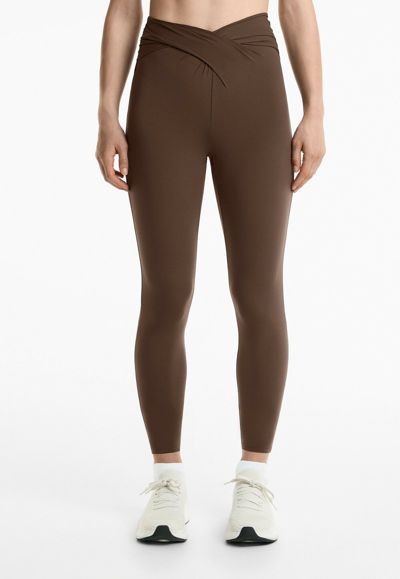 Bruna högmidjade leggings med en vriden design vid midjan. Tillverkade av en elastisk tygkvalitet, de passar nära kroppen.