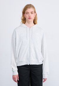 Haut de survêtement zippé gris clair avec une coupe décontractée, une capuche et des poches avant. Tissu doux, poignets et ourlet côtelés, avec un logotype discret sur la poitrine.