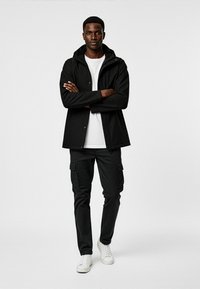 Svart vattentät jacka med huva, kombinerad med svarta cargo-byxor och vita sneakers. Modell står med armarna i kors mot en enkel bakgrund.