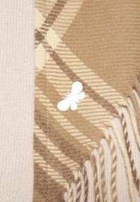 Écharpe à carreaux beige et marron avec des bordures effilochées et une broche insecte blanche stylisée attachée à un pull crème en tricot.