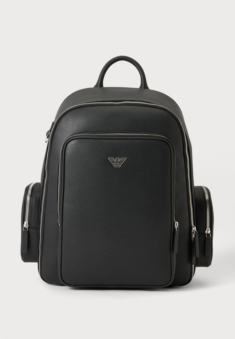 Sac à dos noir avec des fermetures éclair argentées, des poches frontales et latérales, et un petit logo aigle Armani argenté au centre de la face avant.