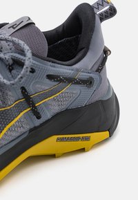Zapato de senderismo gris con parte superior de tejido texturizado, acentos amarillos y suela de goma negra. Presenta un logo PUMAGRIP ATR en el lateral.