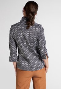 Chemise à motifs noir et blanc avec col montant, manches roulées et fentes sur les côtés, associée à un pantalon marron.