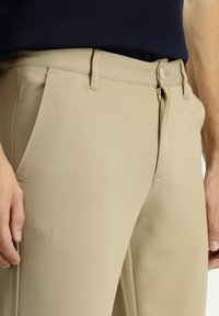 Pantaloni beige con finitura testurizzata, zip frontale e chiusura a bottone. Sono visibili tasche laterali. Adatti per un abbigliamento casual o semi-formale.