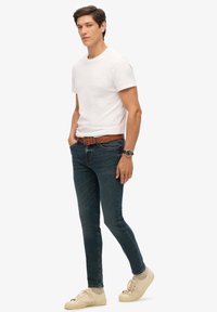 Camiseta blanca de algodón, jeans ajustados de mezclilla azul oscuro, cinturón de cuero marrón con hebilla, zapatillas beige, en pose de pie sobre fondo sencillo.
