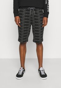 Mężczyzna w czarnych szortach z powtarzającymi się logotypami "Calvin Klein", czarnej bluzie i czarnych skarpetkach Nike z białymi sznurówkami i podeszwami.