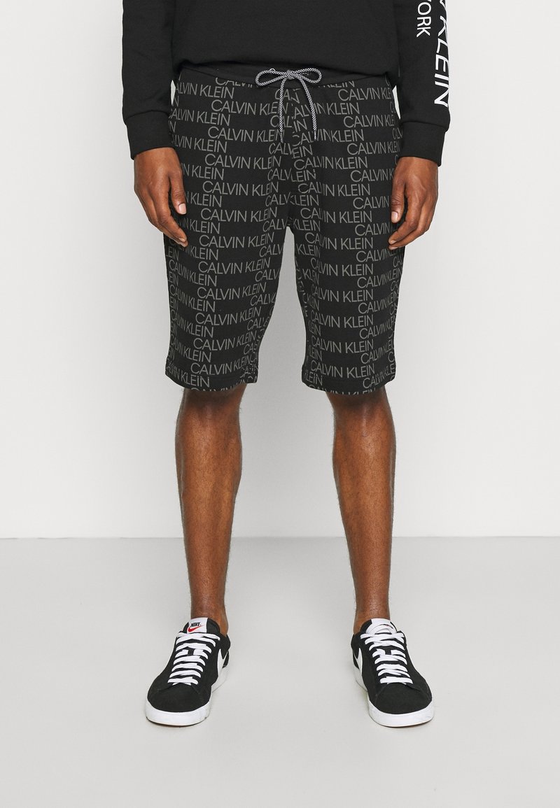 Mężczyzna w czarnych szortach z powtarzającymi się logotypami "Calvin Klein", czarnej bluzie i czarnych skarpetkach Nike z białymi sznurówkami i podeszwami.