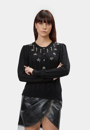 Cardigan nero lavorato a maglia con ricami e applicazioni, presenta un colletto e polsini a coste. Indossato con una gonna in pelle nera con frange.