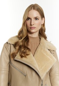 Beige nahkahakki, jossa on pehmeä, vaalea shearling-kaulus. Varustettu vetoketjuyksityiskohdilla ja rakenteellisella, lyhyellä muotoilulla. Pintakuviointi näkyy.