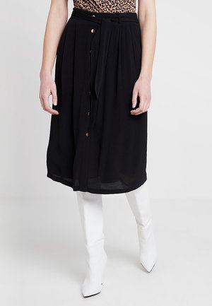 Kobieta ubrana w czarną, midi spódnicę z przodu zapinaną na guziki i przewiązaną paskiem, zestawioną z białymi, wysokimi botkami na szpic oraz topem w panterkę.