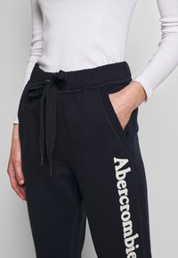 Persoon in zwarte trainingsbroek met "Abercrombie" tekst op het been en een witte longsleeve, hand in de zak tegen een plain achtergrond.