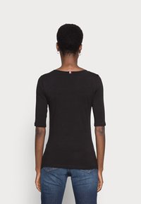 Osoba s krátkými kadeřavými vlasy, oblečená v černém triku s rukávy do lokte a modrých džínách, stojící zády k jednoduchému světlému pozadí.